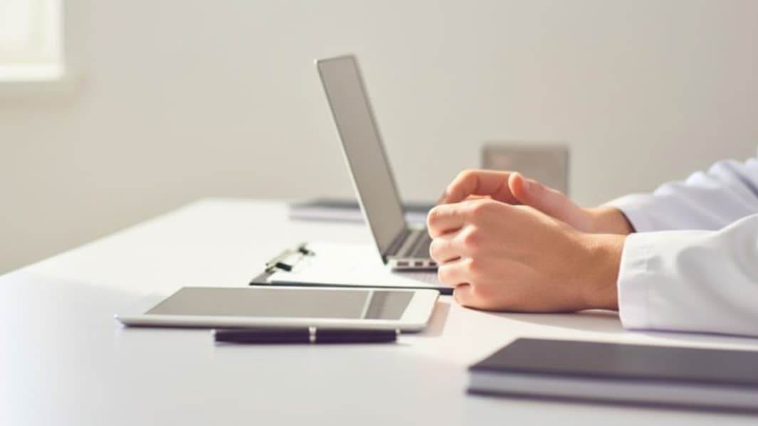 Close-up de uma pessoa em jaleco branco com as mãos entrelaçadas sobre uma mesa com laptop e tablet.